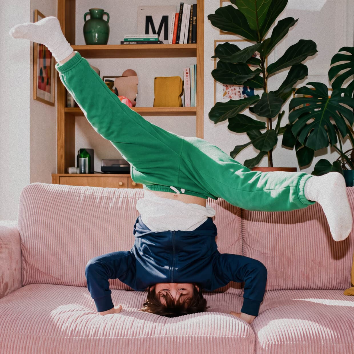 A child in green joggers and a navy top kicks one leg up while balancing on their head on a pink sofa, with a bookshelf and large plant in the background.